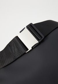 Schwarzer Taschenriemen mit einer silbernen Metallschnalle, aus strukturiertem Stoff und flacher Oberfläche. Enthält Steppnähte entlang des Riemens.