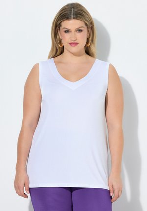 Kvinde med blondt hår iført guldringøreringe, hvid ærmeløs V-hals top og lilla bukser, der står mod en hvid baggrund.