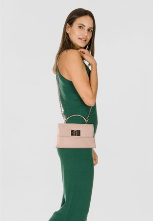 Frau in einem ärmellosen, dunkelgrünen Kleid, die eine hellrosa Handtasche mit Metallverschluss und Kettenriemen über der Schulter trägt.