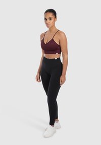 Sujetador deportivo burdeos corto con tirantes finos, combinado con leggings negros de cintura alta y zapatillas deportivas blancas, de pie sobre un fondo neutro.