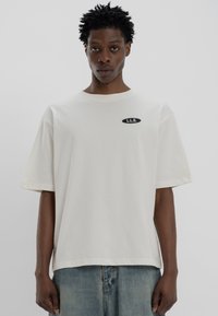 Light beige oversized t-shirt with a round neckline, short sleeves, and a black oval patch featuring white text "L.L.S." stitched on the front.