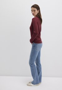 Burgunderfarbener Zopfmuster-Pullover mit gerippten Bündchen, kombiniert mit hellblauen Schlagjeans und beigen Sneakers. Glatte Textur und taillierte Passform.
