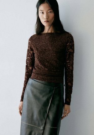 Femme aux longs cheveux foncés porte un haut en dentelle marron à manches longues et une jupe en cuir vert foncé, debout devant un fond clair uni.