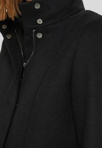 Veste en laine noire avec un col haut, dotée de boutons-pression argentés et d'une fermeture éclair centrale. Le tissu a une texture douce et des lignes épurées.