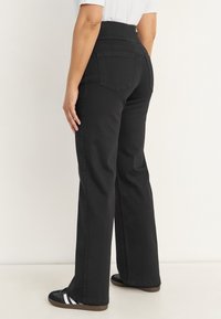 Nainen pukeutuneena korkeavyötäröisiin mustiin leveälahkeisiin farkkuihin ja mustiin tennareihin, joissa on valkoiset raidat, seisoo valkoista taustaa vasten.