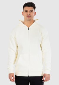 Sweat à capuche zippé couleur crème avec capuche, poches avant et poignets côtelés. Fabriqué en tissu doux et texturé avec un design simple.