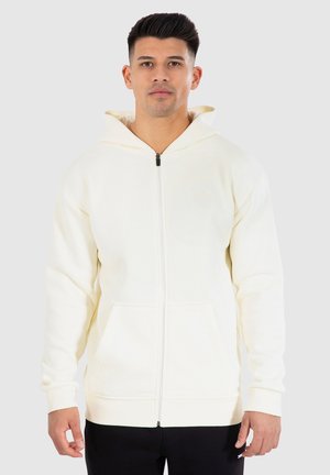 Sweat à capuche zippé couleur crème avec capuche, poches avant et poignets côtelés. Fabriqué en tissu doux et texturé avec un design simple.