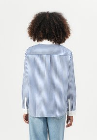 Camicia a righe blu e bianche con maniche lunghe, colletto a punta e vestibilità rilassata. Presenta una piega sul retro e polsini bianchi a contrasto.