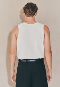 Top bianco senza maniche con texture a coste, abbinato a pantaloni neri e cintura nera. Vista posteriore che mostra i dettagli di spalle e braccia.