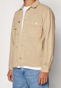 Beige denimjacka med krage, frontknäppning, två bröstfickor med klaffar och en avslappnad passform.