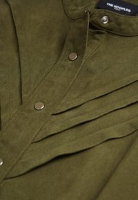 Camisa de pana verde oliva con un diseño de botones y botones de presión de metal, y una textura plisada en la parte delantera.