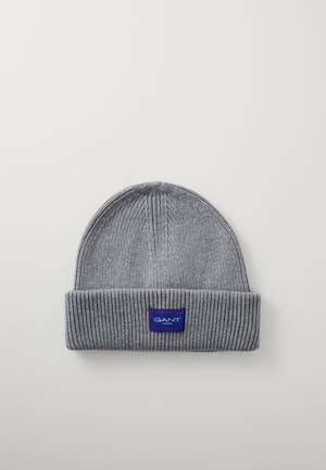 Grijze gebreide beanie met een omgeslagen rand. Voorzien van een blauwe rechthoekige label met witte tekst aan de voorkant. Zachte textuur en geribbelde ontwerp.