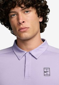 Lys lilla Nike polo-t-shirt med en struktureret stof, der har en klassisk krave, to knapper og et sort logo på venstre bryst.