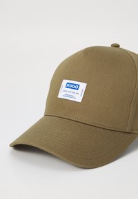 Casquette de baseball khaki en tissu texturé, avec un patch logo rectangulaire bleu et blanc à l'avant. Visière incurvée, design simple.