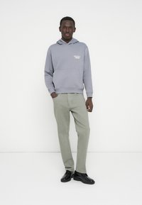 Γκρι φούτερ με κουκούλα, μπροστινή τσέπη και λογότυπο, συνδυασμένο με ανοιχτό πράσινο παντελόνι και μαύρα παπούτσια. Ομαλή υφή, casual σχεδιασμός.