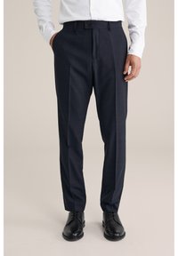 Uomo che indossa pantaloni eleganti su misura blu navy scuro, scarpe eleganti nere e una camicia bianca a maniche lunghe con bottoni, in piedi con una mano in tasca.