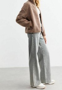 Ruskea, pörröinen fleece-takki, jossa on vetoketjusuljenta edessä, yhdistetty vaaleanharmaisiin leveälahkeisiin farkkuihin ja valkoisiin tennareihin.