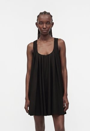 Kobieta z warkoczami, ubrana w czarną plisowaną sukienkę bez rękawów, stojąca na jednolitym białym tle, zwrócona przodem.