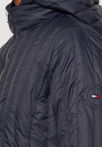 Chaqueta acolchada azul marino con collar alto, cremallera plateada visible y parche del logo de Tommy Hilfiger en la parte superior de la manga.