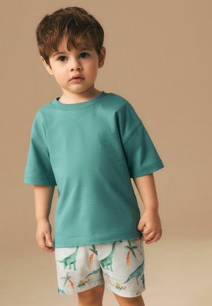 Fiatal fiú rövid barna hajjal, türkiz pólót és fehér rövidnadrágot visel, amely zöld dinoszaurusz és pálmafás mintákkal díszített, bézs háttér előtt áll.