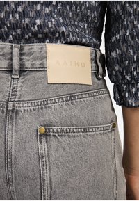 Grijze denim jeans met een leren label met "AAIKO," zichtbare stiksels en messing studs op de achterzakken. Gestructureerde stof.