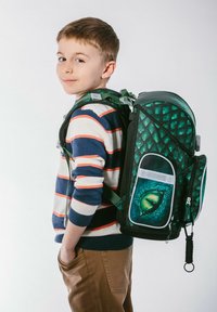 Plecak w zielonym motywie smoka, z teksturowanymi łuskami, wieloma przegródkami oraz żywym detalem w postaci oka. Dziecko noszące sweter w paski i beżowe spodnie.