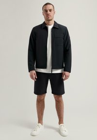 Giacca nera con zip, colletto e tasca sul petto, abbinata a pantaloni corti neri. Modello che indossa una maglietta bianca e sneakers bianche.