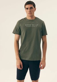 Zielona bawełniana koszulka z nadrukiem "BUDŹ MNIE" oraz "HENDERSON APPAREL CO." w białym kolorze z przodu. Okrągły dekolt, krótkie rękawy, luźny krój.