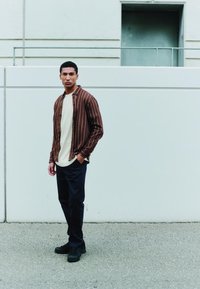 Bruin gestreept flanellen overhemd gedragen over een crèmekleurig T-shirt met ronde hals; gecombineerd met donkere broek en zwarte veterlaarzen. Effen kleuren en texturen aanwezig.