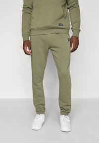 Olivu zaļa kokvilnas sporta bikses ar šauru piegriezumu, elastīgu jostu un divām kabatām, apvienotas ar baltām sporta apavām.