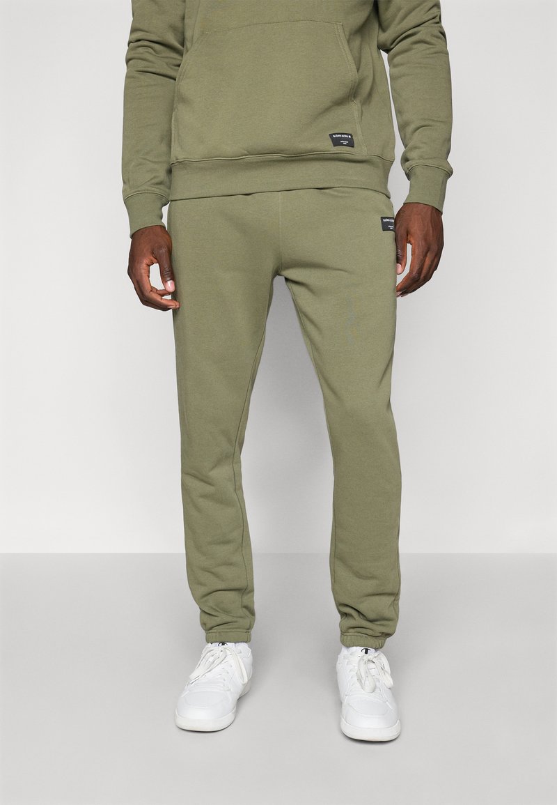 Olivu zaļa kokvilnas sporta bikses ar šauru piegriezumu, elastīgu jostu un divām kabatām, apvienotas ar baltām sporta apavām.