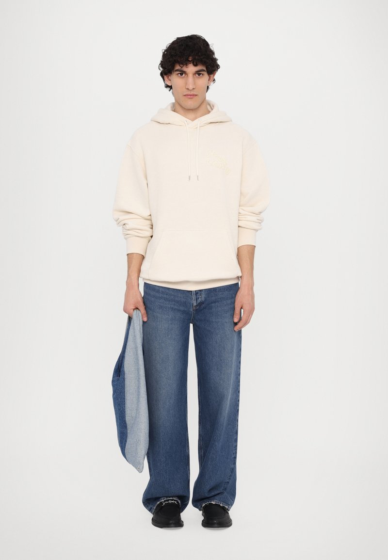 Beige Kapuzenpullover mit Fronttasche und gesticktem Logo, kombiniert mit weit geschnittenen blauen Jeans und schwarzen Schuhen. Hält eine blaue Einkaufstasche.