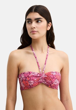 Młoda kobieta o ciemnych włosach, ubrana w różowo-czerwony bikini typu bandeau w liściasty wzór z ramiączkiem na szyję, patrząca lekko na bok.