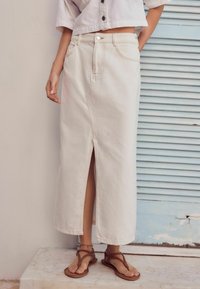 Witte denim maxirok met een zijsplit, hoge taille en zichtbare stiksels. Gecombineerd met bruine platte sandalen op een lichte ondergrond.