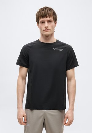 Mladý muž stojaci čelom k fotografovi, oblečený v čiernom tričku s krátkym rukávom a malým bielym nápisom na hrudi, a v béžových nohaviciach.