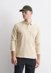 Bež polo majica s dolgimi rokavi in ovratnikom, z diskretno logo in rebrastimi manšetami, v kombinaciji z temnimi hlačami in zeleno kapo.