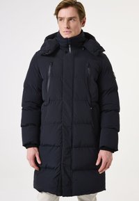 Abrigo largo negro tipo puff con capucha, bolsillos con cremallera, cierre frontal con cremallera y textura acolchada. Presenta puños elásticos y un ajuste entallado.