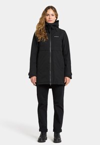 Black waterproof jacket with a zip closure, adjustable hood, and two side pockets. Worn over black pants and sturdy black boots.