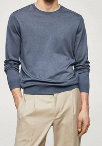 Mann in einem taillierten blauen Langarmpullover, der in beigen, gefalteten Hosen steckt und mit einer Hand in der Tasche steht, vor einem schlichten Hintergrund.