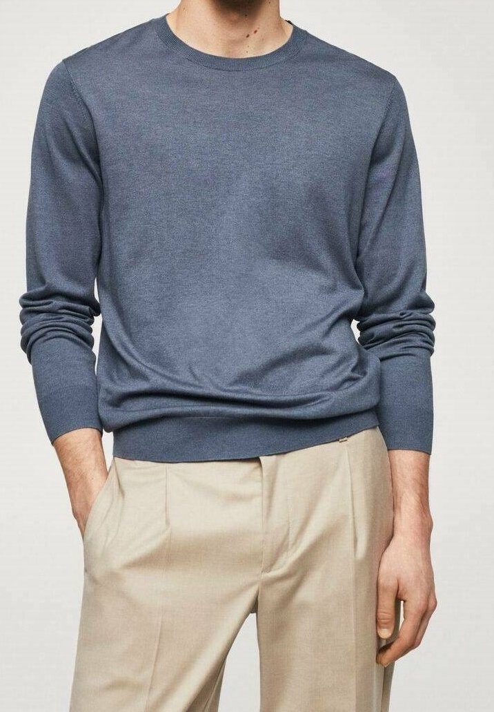 Mann in einem taillierten blauen Langarmpullover, der in beigen, gefalteten Hosen steckt und mit einer Hand in der Tasche steht, vor einem schlichten Hintergrund.