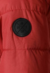 Giacca a vento rossa con un patch rotondo in gomma nera con marcature in rilievo. Il tessuto è liscio, con cuciture visibili e una superficie testurizzata.