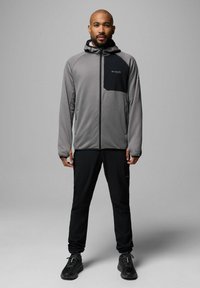 Szara lekka kurtka z czarnymi akcentami, zapinana na zamek z przodu i z kapturem, zestawiona z czarnymi spodniami i czarnymi trampkami. Gładka faktura i sportowy krój.