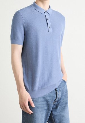 Man draagt een lichtblauw gebreid poloshirt met korte mouwen en een blauwe spijkerbroek, met één hand in de zak en de andere langs zijn lichaam.