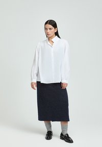 White long-sleeve button-up shirt with a high collar, paired with a dark textured skirt and gray socks, worn with black loafers.