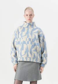 Fleece pullover in lichtblauw en roomkleur met geometrische patronen; heeft een hoge kraag en een halve rits; zachte textuur; oversized pasvorm.