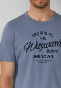 Man in een lichtblauw T-shirt met zwarte tekst "Escape to the Wayward Spirit Unknown S.Oliver Unbound Since 1969."