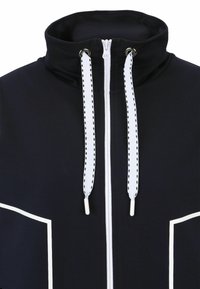 Marinelint zip-upjack met hoge kraag, contrasterende witte stiksels en trekkoorden. Heeft grote witte accentzakken aan de voorkant.