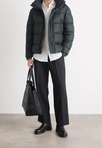 Uomo che indossa un piumino nero, una camicia bianca, pantaloni neri e stivali neri, mentre tiene una grande borsa tote nera, in piedi su un pavimento grigio.