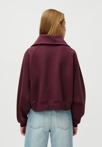 Woman seen from behind wearing a loose maroon sweatshirt with a wide collar and light blue high-waisted jeans against a white background.
