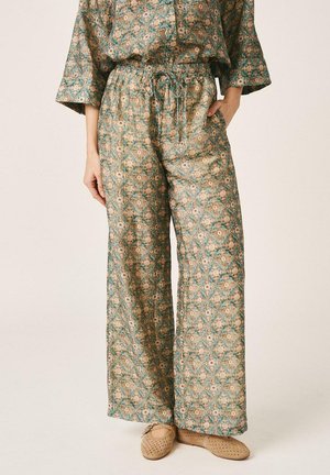 Mujer que lleva pantalones sueltos de cintura alta con un patrón floral verde y beige, combinados con un top a juego y zapatos beige perforados sin cordones.
